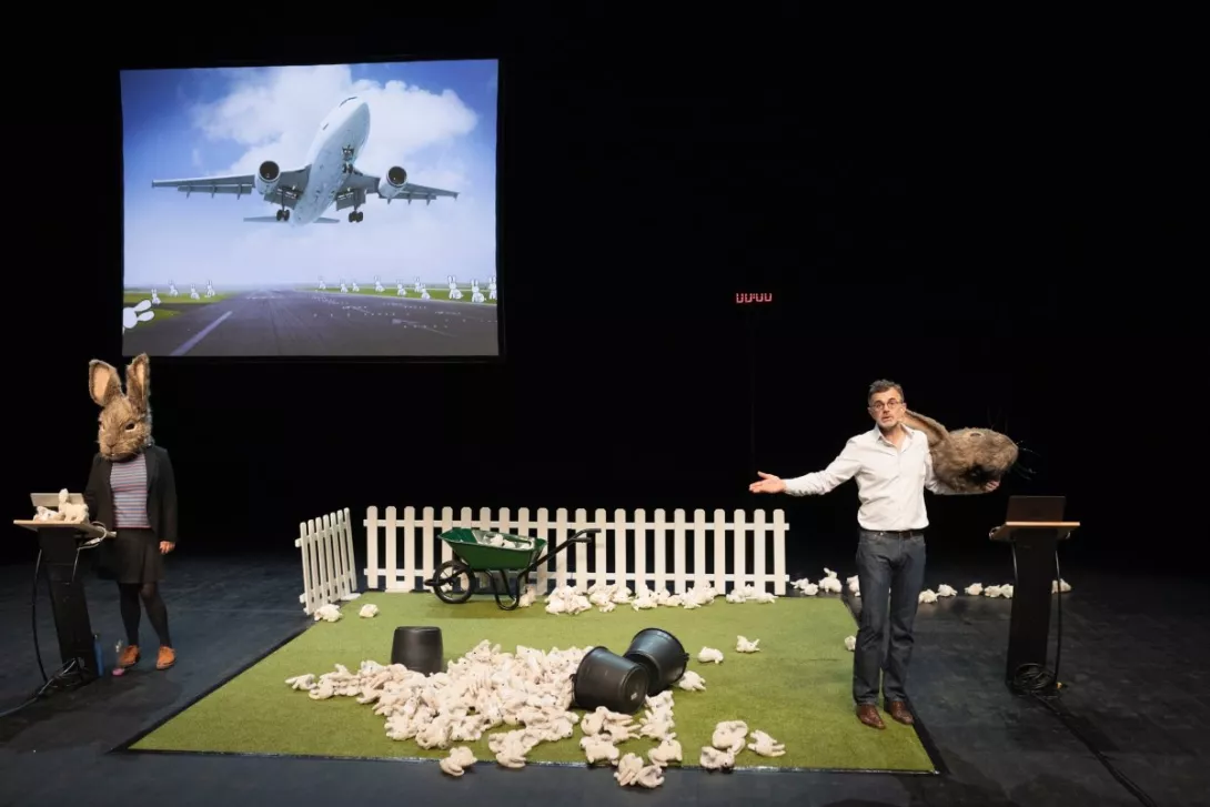 Une scène du spectacle Le problème lapin : deux comédiens de chaque côté de la scène avec des masques de lapin et un tas de lapin en peluche au milieu sur une fausse pelouse verte. Un écran au fond avec un avion qui décole. 